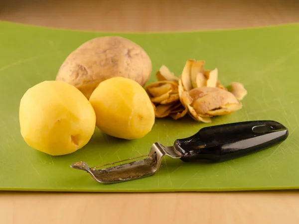 Three potatoes and peeler on used green board — Stock Photo, Image