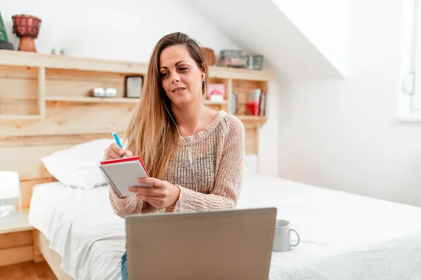 Dama casual tomando notas mientras está de guardia desde su habitación usando una computadora con auriculares, mujer de negocios tomando notas a través de la reunión de conferencia Vedio desde un ordenador portátil en el dormitorio — Foto de Stock