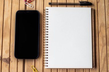 Boş Kalemleri Ahşap Masa 'nın üzerinde olan Boş Bir Günlük. Boş Spiral Not Defteri, masaya yerleştirilmiş balon kalemli bir telefonun yanında..