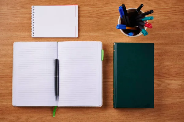 Nyílt lapos jegyzetfüzet egy halom tollal az asztal tetején. Egyszerű üres soros jegyzettömbök mellett egy tulajdonosa színes golyóstollak egy íróasztalon. — Stock Fotó