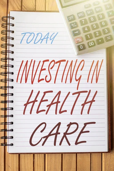Hand writing sign Investing In Health Care. Business idea put money on maintenance or improvement of health Blank Open Spiral Notebook With A Calculator Placed On Table.