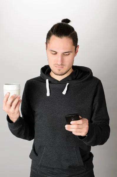 Joven alegre usando un teléfono inteligente y sosteniendo una taza de bebida . — Foto de Stock