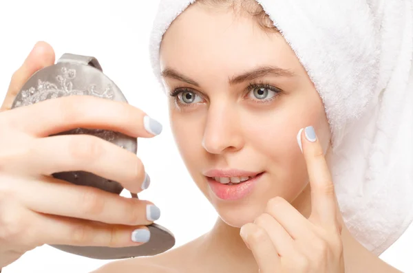 Bella giovane donna che applica una crema sul suo viso guardando specchio isolato su sfondo bianco — Foto Stock