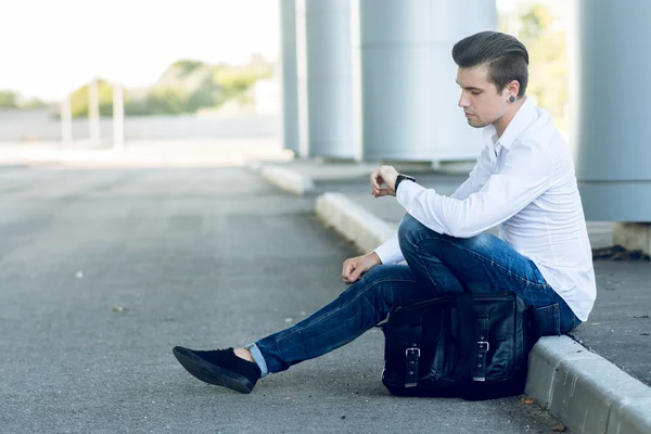 Joven guapo usando teléfono inteligente mientras está sentado cerca de la carretera — Foto de Stock