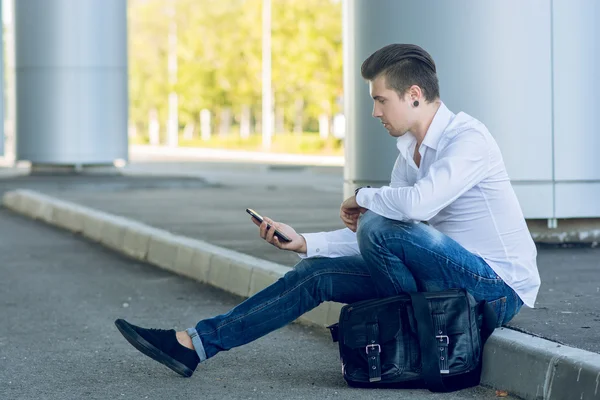 Schöner junger Mann benutzt Smartphone, während er in der Nähe der Straße sitzt — Stockfoto