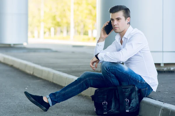 Joven guapo hablando teléfono inteligente mientras está sentado cerca de la carretera — Foto de Stock