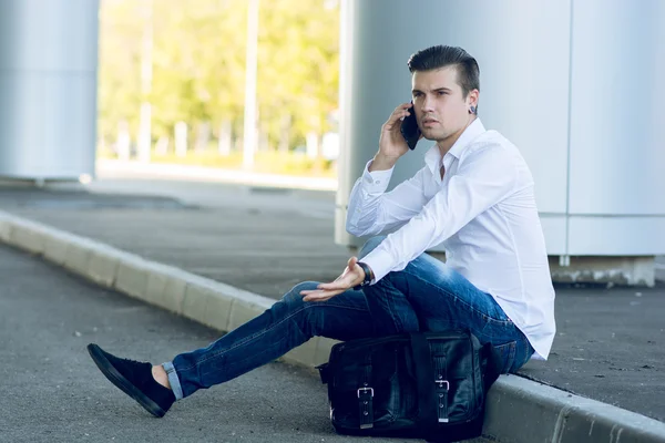 Joven guapo hablando teléfono inteligente mientras está sentado cerca de la carretera — Foto de Stock
