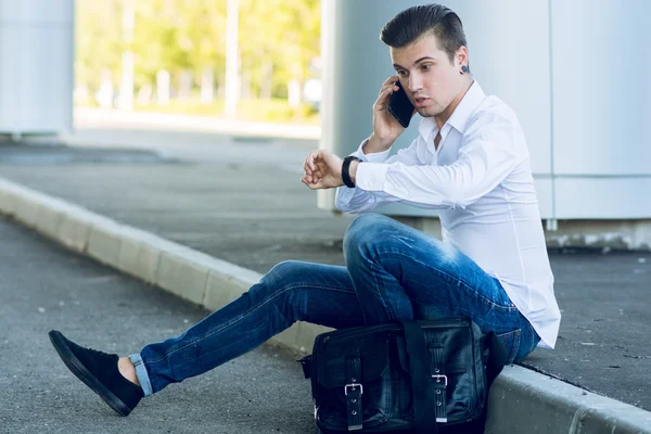 Joven guapo hablando teléfono inteligente mientras está sentado cerca de la carretera — Foto de Stock