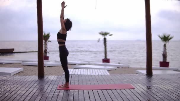 Mujer joven está haciendo yoga en el suelo de madera en la playa, la chica realiza puestos de yoga y elementos — Vídeos de Stock