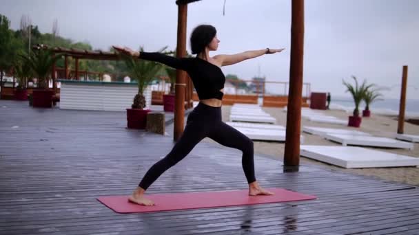 Chica está haciendo yoga en la estera en la playa, la chica realiza puestos de yoga y elementos - guerrero — Vídeos de Stock