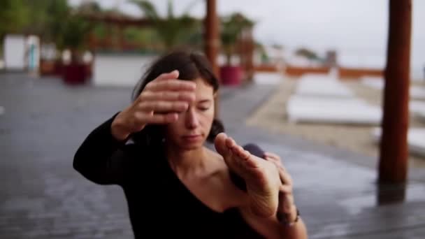 Chica de negro está haciendo yoga en el suelo de madera en la playa, la chica realiza elemento de yoga - doblar la pierna detrás del hombro — Vídeos de Stock