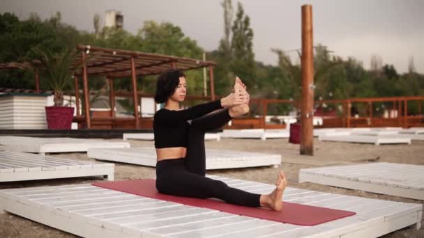 Frau praktiziert Yoga sitzend auf Matte am Strand und führt Yoga-Asanas und Elemente durch oder streckt die Beine — Stockvideo