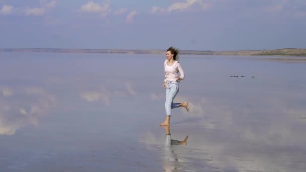 Heureuse femme aux cheveux longs dans casual est en cours d'exécution sur le sable peu profond du lac avec miroir de réflexion de surface — Video