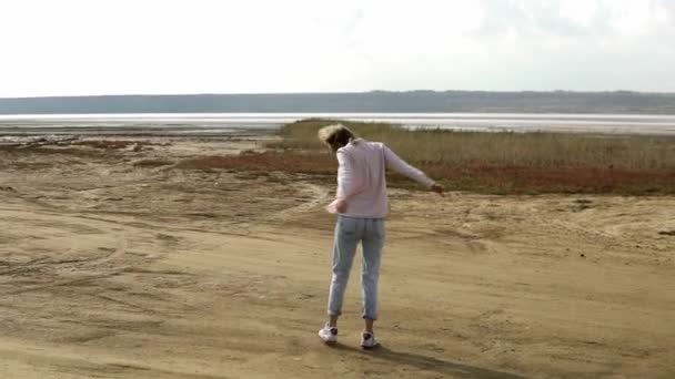 Zorgeloze jonge vrouw danst alleen staan op zand strand met rustige meer op de achtergrond — Stockvideo