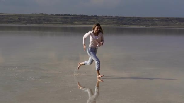 Felice donna dai capelli lunghi in casual è in esecuzione su sabbia superficiale del lago in estate e sorridente — Video Stock