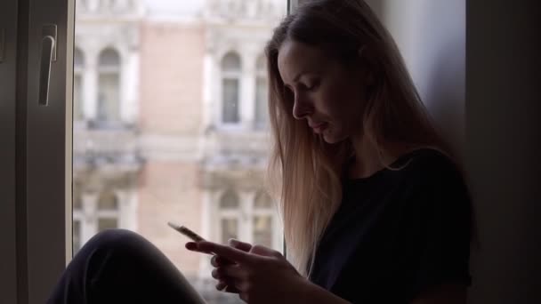Mujer joven sentada en el alféizar de la ventana y usando teléfono inteligente — Vídeo de stock