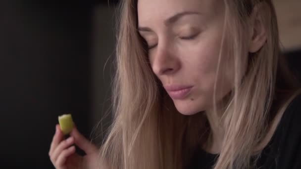 Mujer joven rubia de pie en la cocina comiendo manzana en el interior de casa — Vídeo de stock