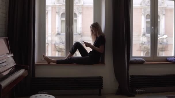 Mujer rubia sentada en el alféizar de la ventana y el uso de teléfono inteligente en la sala de estar con piano — Vídeos de Stock