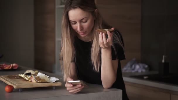 Portret van een vrouw die brood eet met boter als ontbijt en haar smartphone controleert in de keuken — Stockvideo