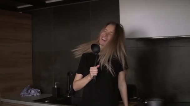 En glad kvinna med en slev i ett modernt kök låtsas sjunga — Stockvideo