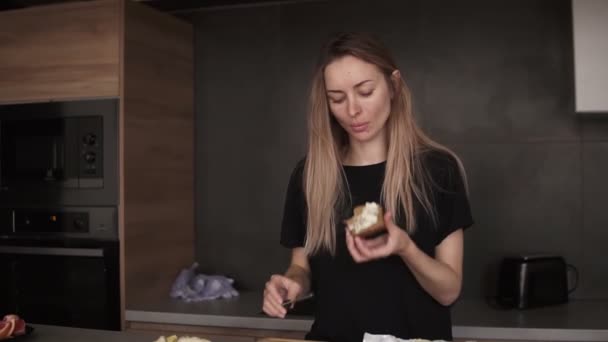 Mulher loira comendo pão com manteiga para o café da manhã, verificando seu smartphone na cozinha — Vídeo de Stock