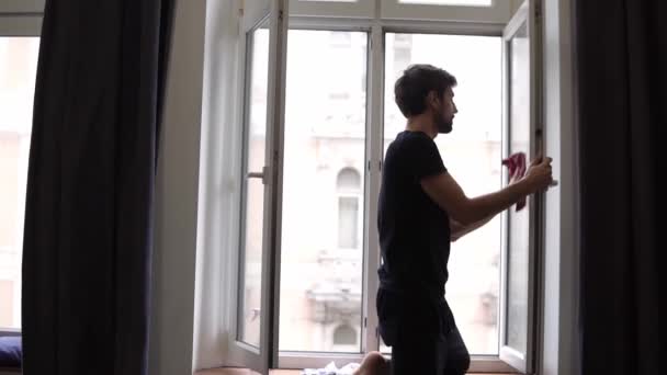 Szakállas férfi ablakot tisztít, felületet tisztít, rongyot használ, modern lakásban áll. — Stock videók