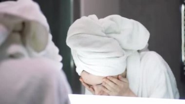 Kafasında havlu olan bir kadın yüzünü suyla yıkar ve aynaya bakar.
