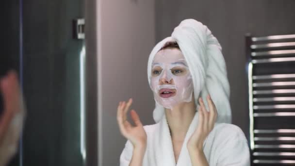 Portré egy nő fürdőköpenyben fehér maszk hidratáló az arcon, nézi a tükröt