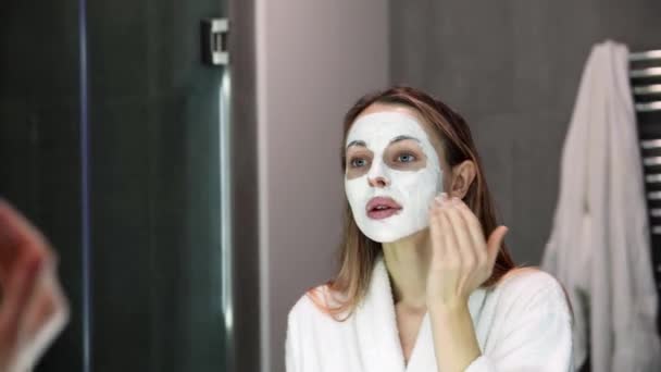 Gyönyörű nő fürdőköpenyben tesz fehér maszk hidratáló ujjak, lassú — Stock videók