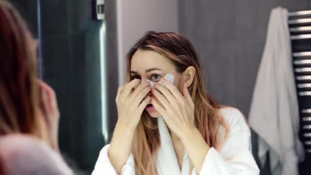 Молода жінка в халаті наносить білі плями очей, дивлячись на дзеркало — стокове відео
