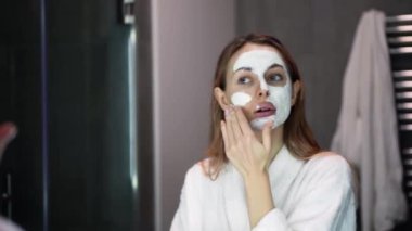 Bornozlu sarışın kadın, parmak ve ağır çekimde nemlendirici için beyaz maske takıyor.