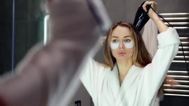 Señora con pelo claro y parches para los ojos mirando su reflejo en el espejo del baño y secándose el cabello con secador de pelo — Vídeo de stock