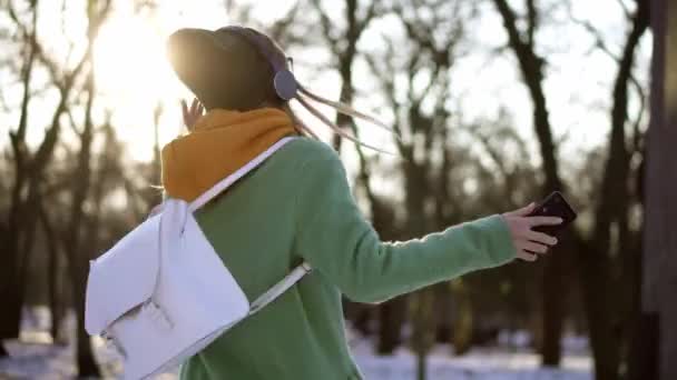 Mulher feliz dança no parque de inverno, ouvindo música no telefone usando fones de ouvido — Vídeo de Stock