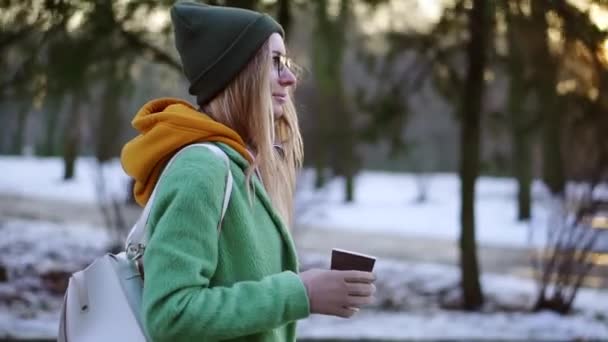 Kobieta spacerując z gorącym napojem przez zimowy park miejski lub las, cieszyć się pogodą, widok z boku — Wideo stockowe