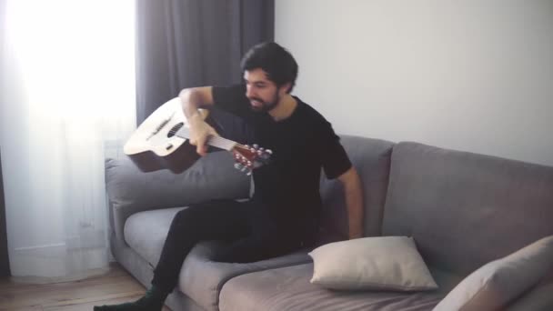 Seorang pemuda duduk di sofa dan bermain gitar — Stok Video