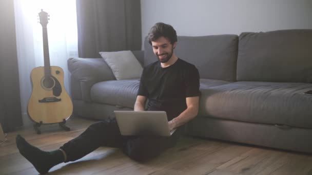 Sonriente hombre sentado en el suelo, mientras que las videollamadas utilizando el ordenador portátil en casa — Vídeos de Stock