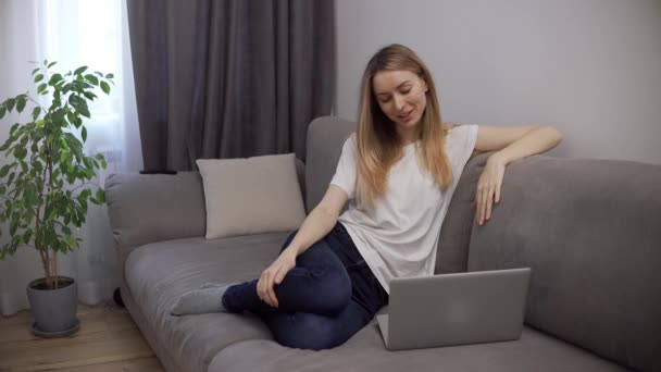 Szőke nő pihen a kanapén, miközben videohívás segítségével laptop otthon — Stock videók