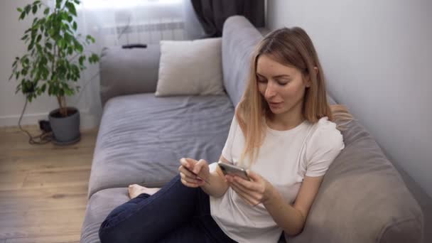 Blonde woman sitting on a sofa, pays for purchases online with a credit card and phone home — Stock Video