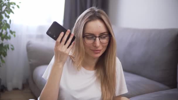 Chica rubia usando asistente virtual en el teléfono móvil, escuchando mensajes de audio y sonriendo — Vídeo de stock