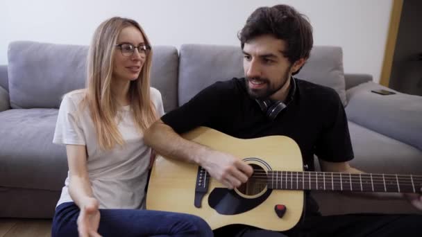 Homem de fones de ouvido toca guitarra em casa, cantando alegremente com sua namorada — Vídeo de Stock