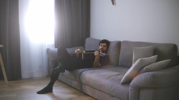Seorang pemuda santai di sofa dan bermain gitar — Stok Video