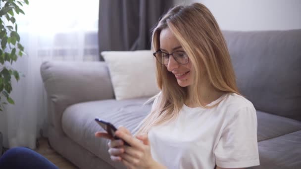 Mooie vrouw chatten op haar telefoon en glimlachen, zitten op de vloer van de woonkamer tijdens quarantaine tijd — Stockvideo