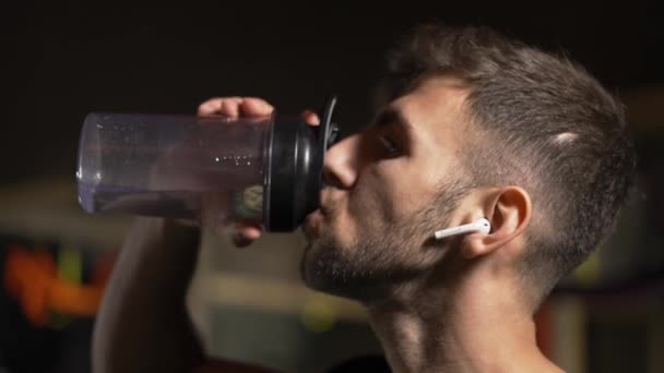 Uomo duro che beve acqua dalla bottiglia nella palestra buia, da vicino — Video Stock
