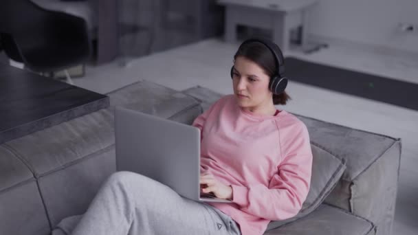 Mujer trabajando con ordenador portátil y escuchando música por auriculares en un sofá — Vídeos de Stock