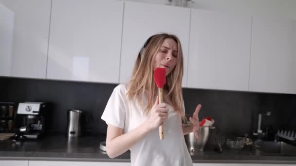 Молода жінка в навушниках танцює і прикидається співом на кухні вдома — стокове відео