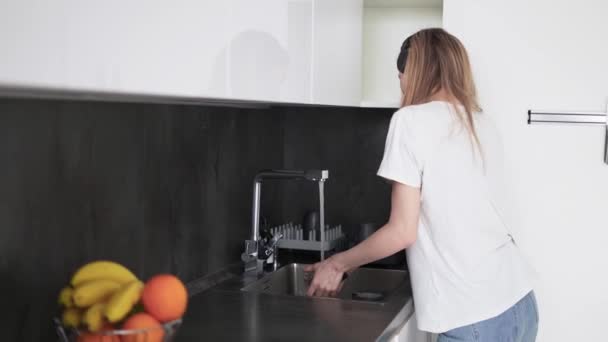 Стройная молодая девушка в наушниках танцует и моет посуду на кухне — стоковое видео