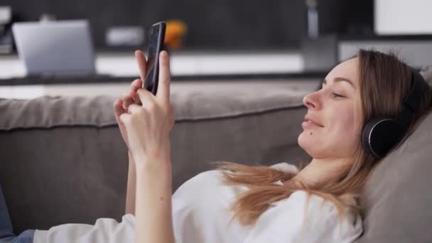 Nő élvezi szabadidejét laiyng egy kanapén zenével és okostelefon — Stock videók