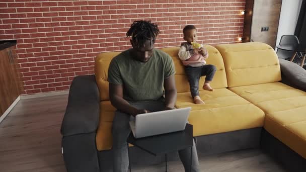 Занятой молодой африканский отец делает внештатную работу на ноутбуке и нянчится с маленьким ребенком дома — стоковое видео
