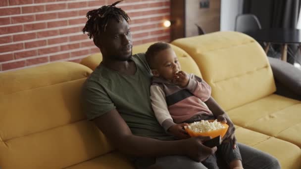 Africký americký otec tráví čas s malým synem sedí na gauči jíst popcorn sledování televize — Stock video