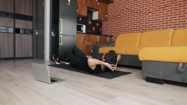 Человек тренируется на коврике с ноутбуком дома, делает велосипедные упражнения на спине — стоковое видео
